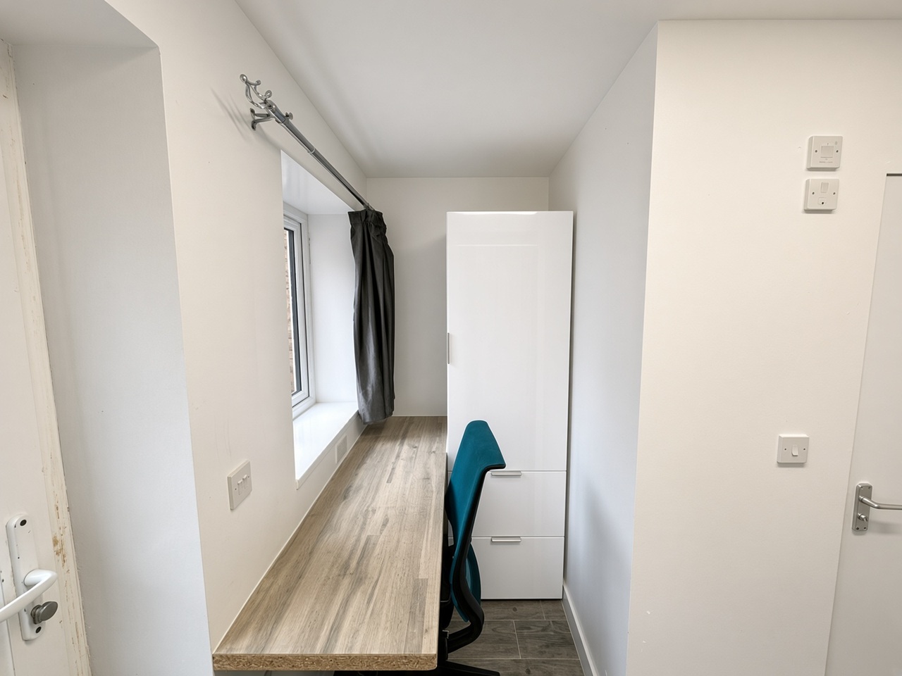 Narrow bedroom with a built-in light-wood desk, teal chair, and white wardrobe