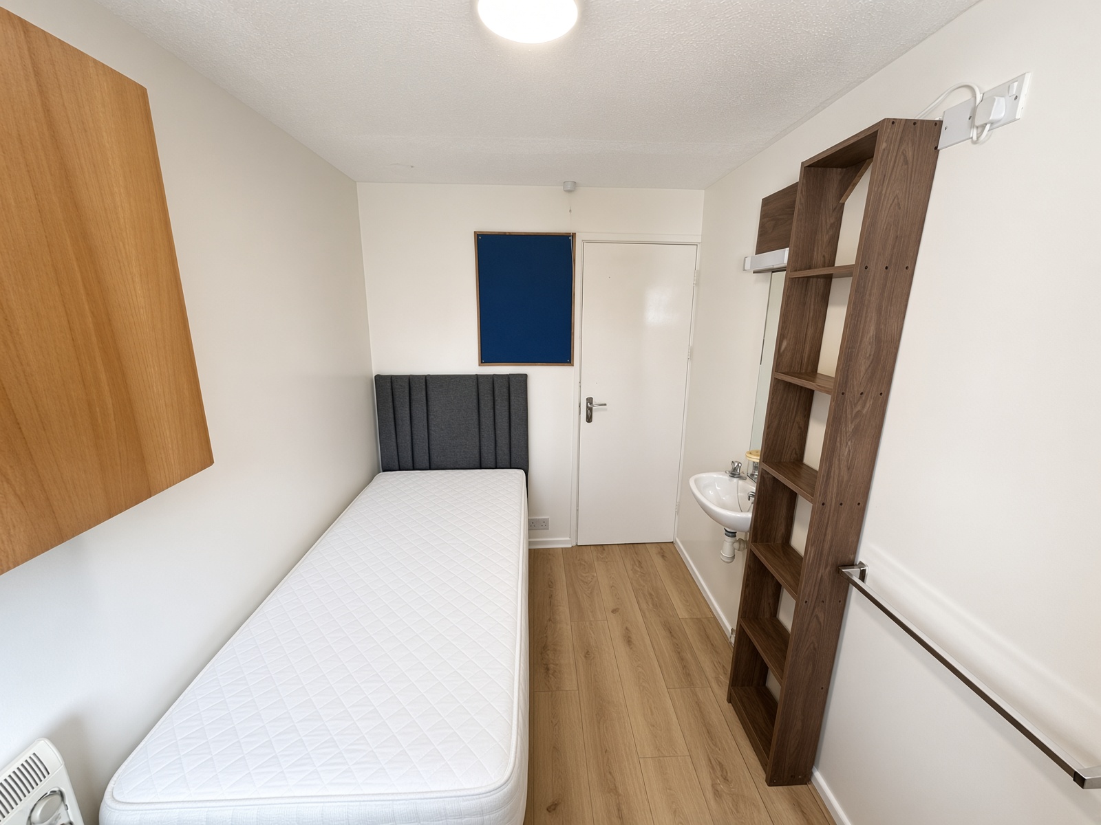 Narrow bedroom with a grey headboard, a washbasin, and a blue notice board