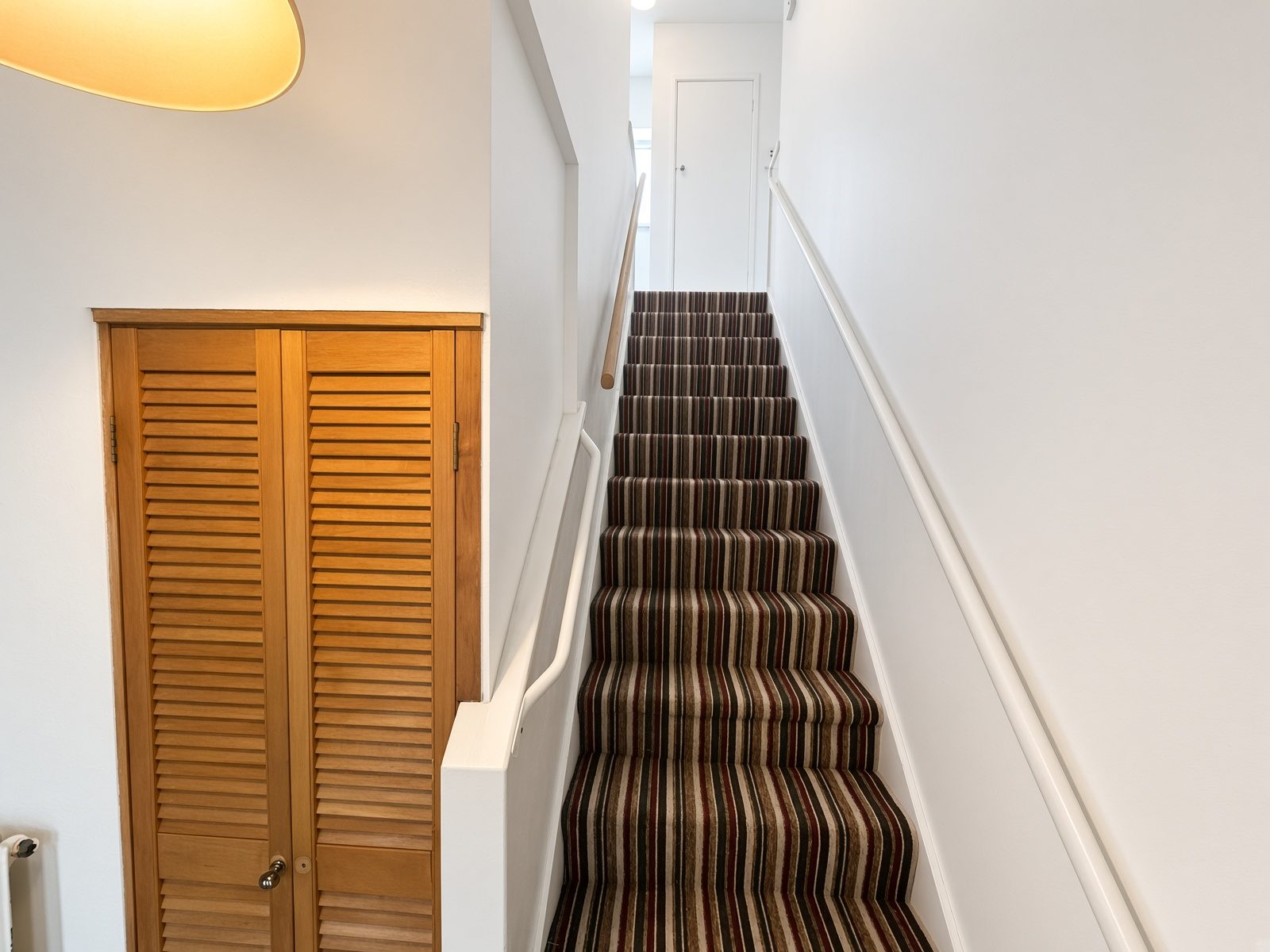 Staircase going up, with striped carpet and a louvered cupboard